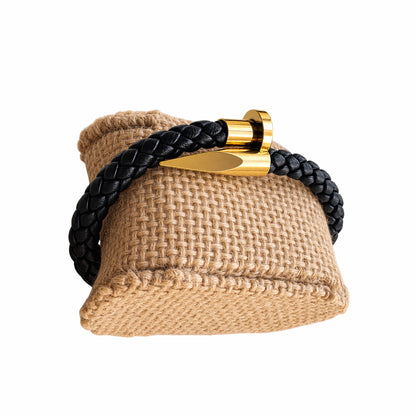 A black braided leather bracelet with a polished gold metal spike nail clasp displayed on a woven tan pillow against a white background