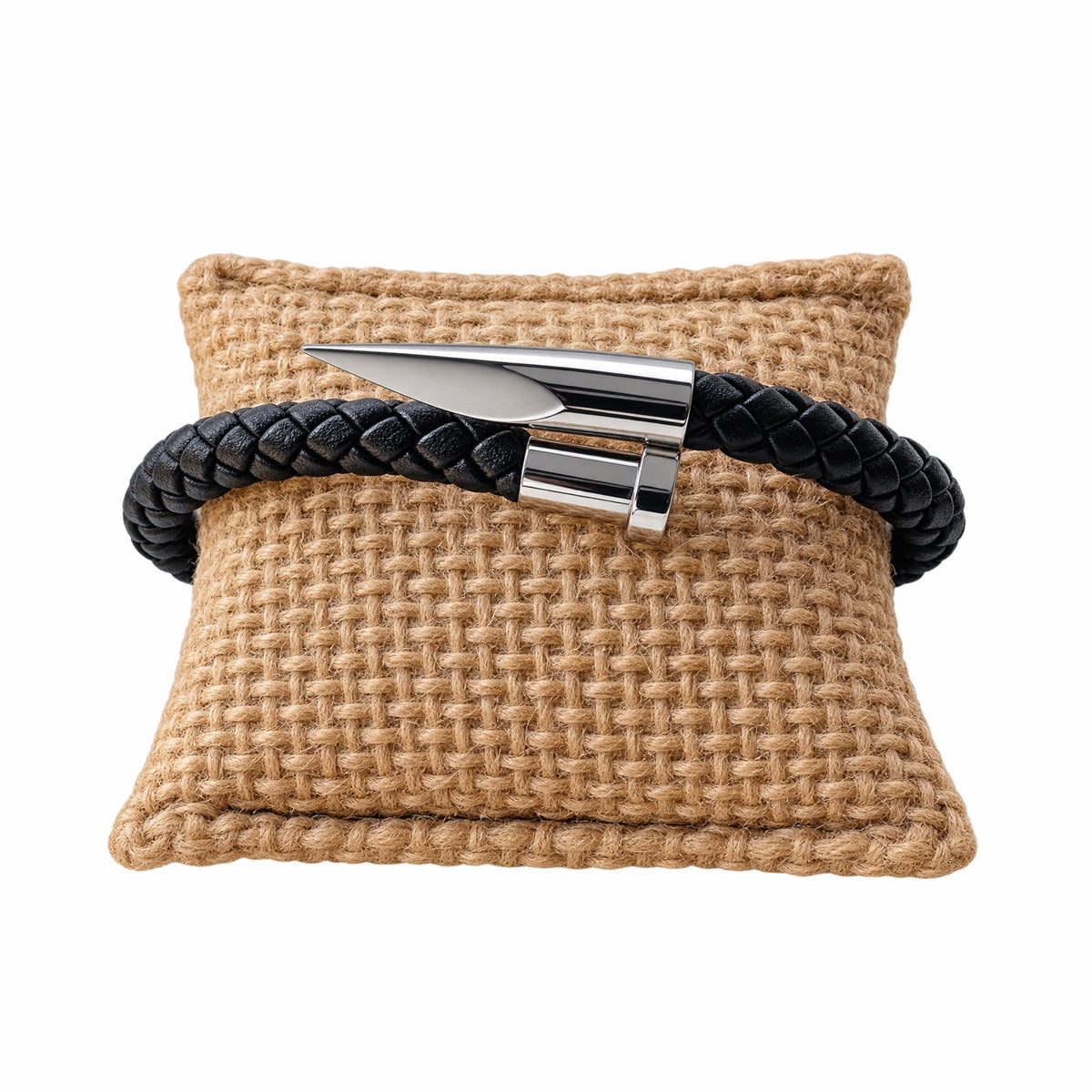 A black braided leather bracelet with a polished silver metal spike nail clasp, displayed on a woven tan pillow against a white background.