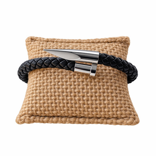 A black braided leather bracelet with a polished silver metal spike nail clasp, displayed on a woven tan pillow against a white background.