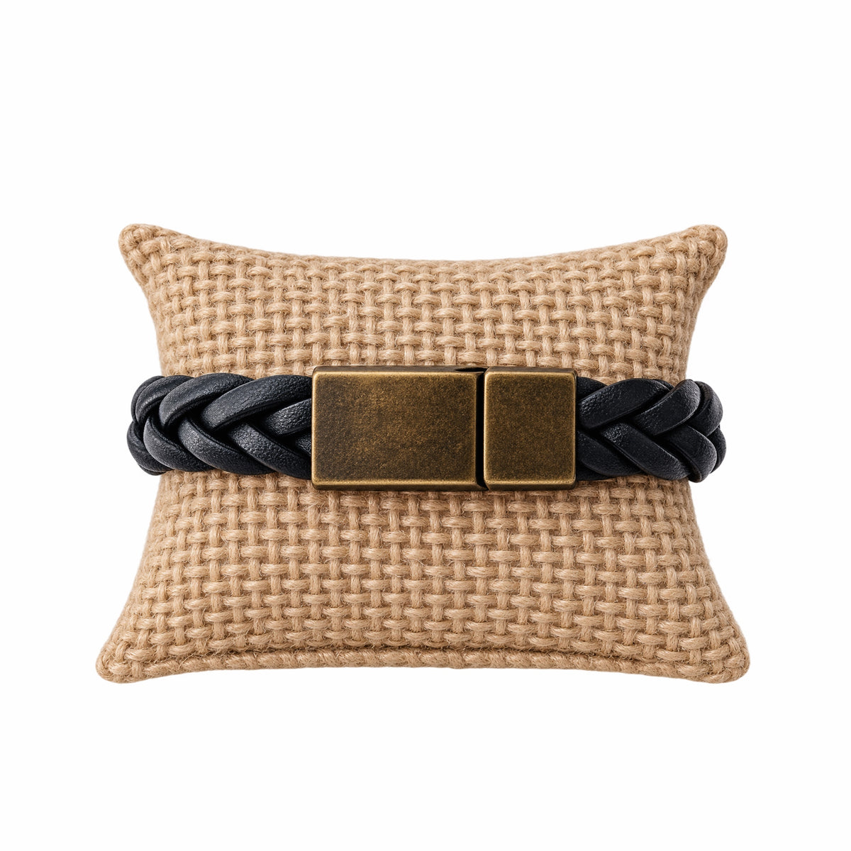 A black braided leather bracelet with a rectangular antique bronze clasp, displayed on a woven beige pillow against a white background.