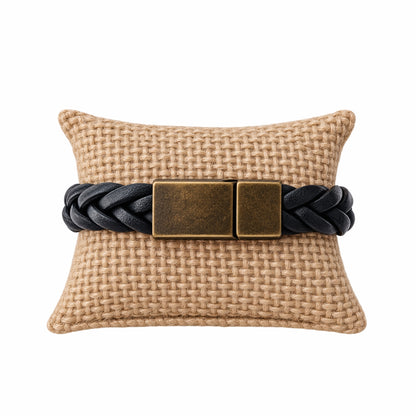 A black braided leather bracelet with a rectangular antique bronze clasp, displayed on a woven beige pillow against a white background.