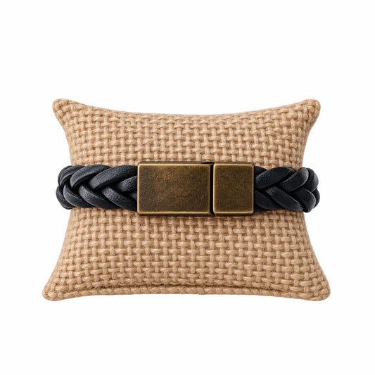 A black braided leather bracelet with a rectangular antique bronze clasp, displayed on a woven beige pillow against a white background.
