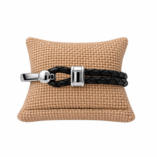 A black braided leather bracelet with polished silver hardware, displayed on a tan woven cushion against a white background.