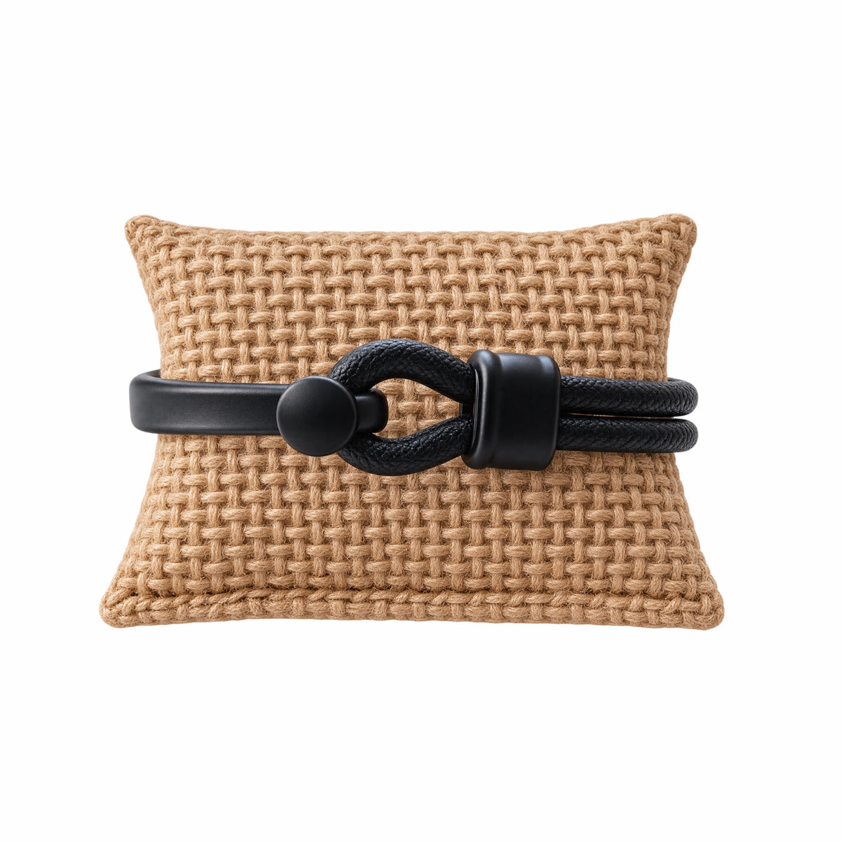 A black leather bracelet with matte black hardware displayed on a tan woven cushion against a white background