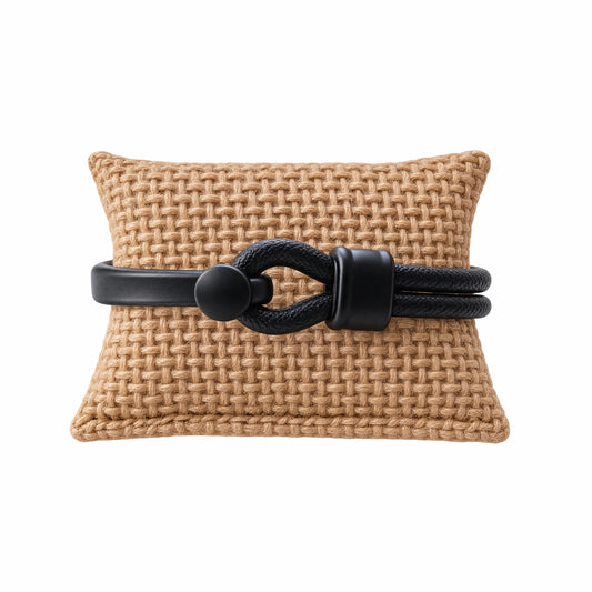 A black leather bracelet with matte black hardware displayed on a tan woven cushion against a white background