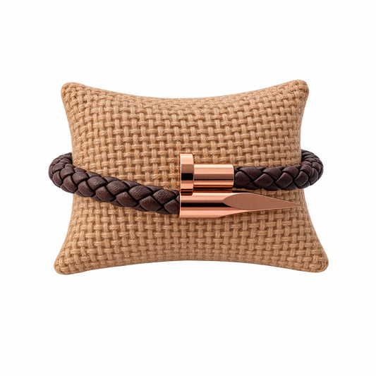 A brown braided leather bracelet with a polished rose gold metal spike nail clasp displayed on a woven tan pillow against a white background