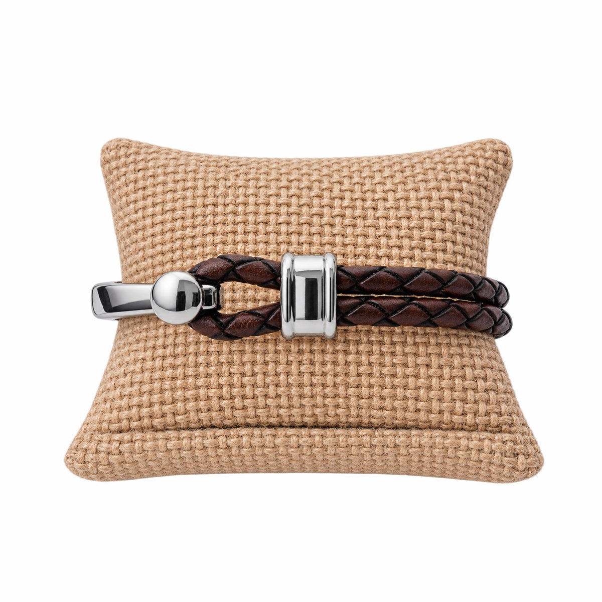 A brown braided leather bracelet with polished silver hardware displayed on a tan woven cushion against a white background