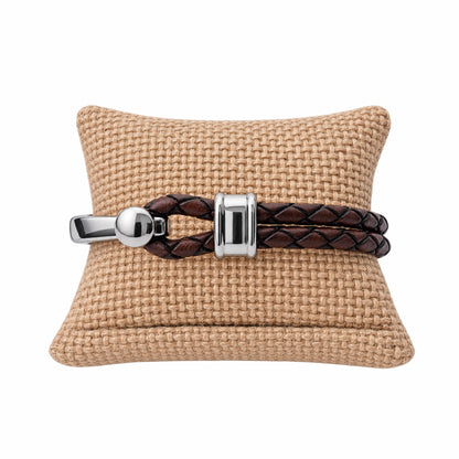 A brown braided leather bracelet with polished silver hardware displayed on a tan woven cushion against a white background