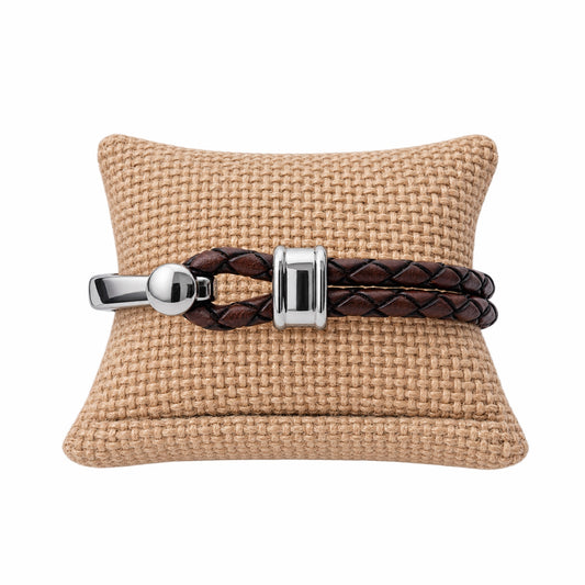 A brown braided leather bracelet with polished silver hardware displayed on a tan woven cushion against a white background