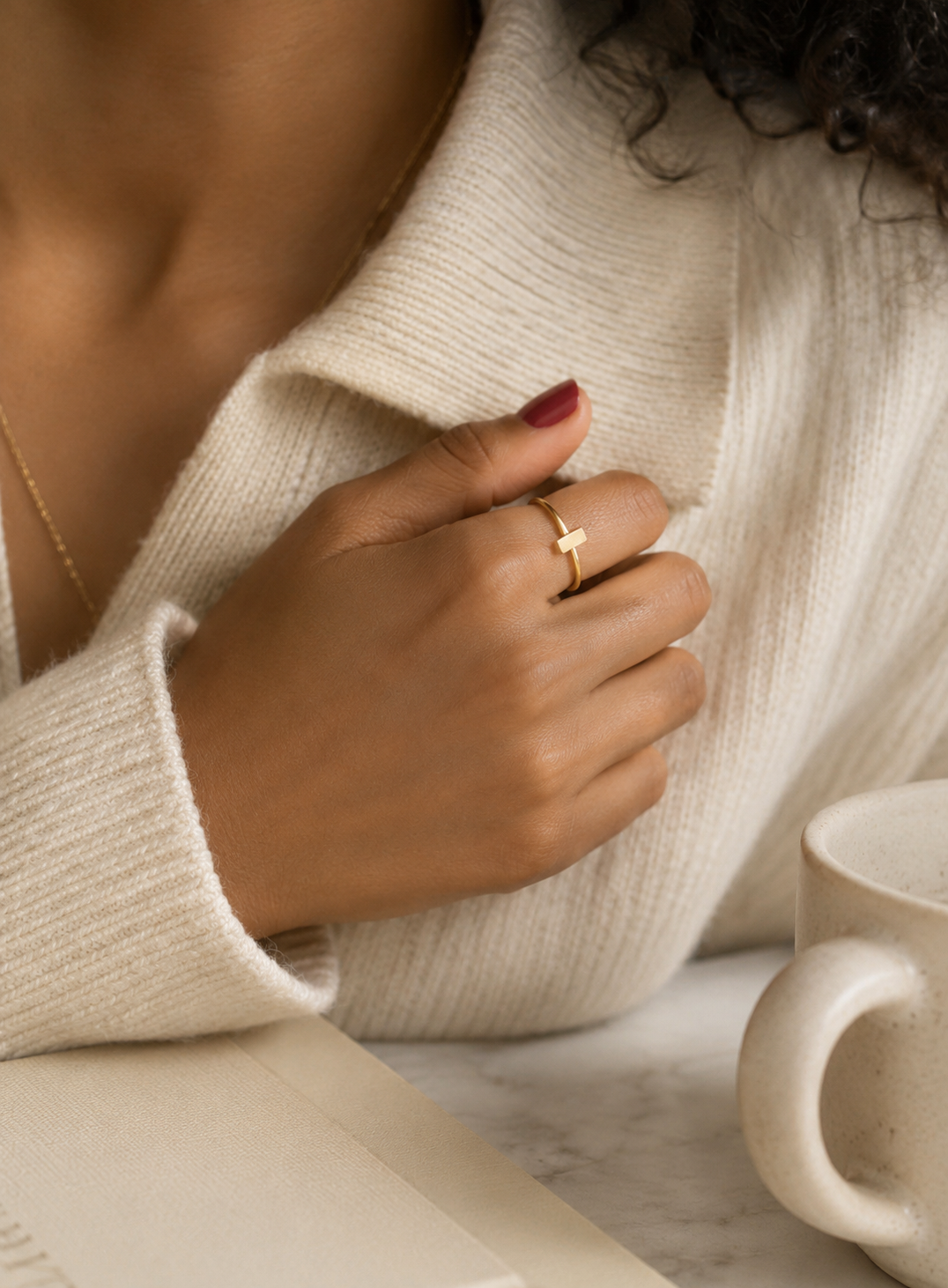 A close-up lifestyle shot of a person wearing a delicate gold bar ring on their ring finger, dressed in a cream-colored knit sweater, with a white ceramic mug on a marble surface in the background.
