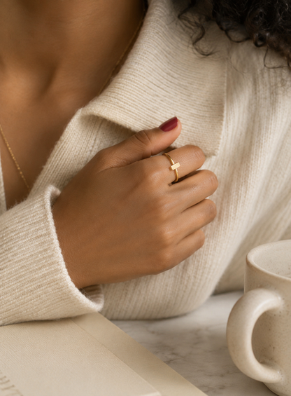 A close-up lifestyle shot of a person wearing a delicate gold bar ring on their ring finger, dressed in a cream-colored knit sweater, with a white ceramic mug on a marble surface in the background.