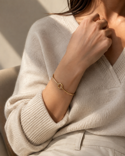 A close-up lifestyle shot of a woman wearing a delicate gold bangle bracelet with a small hexagon detail, paired with a cream-colored ribbed knit sweater.