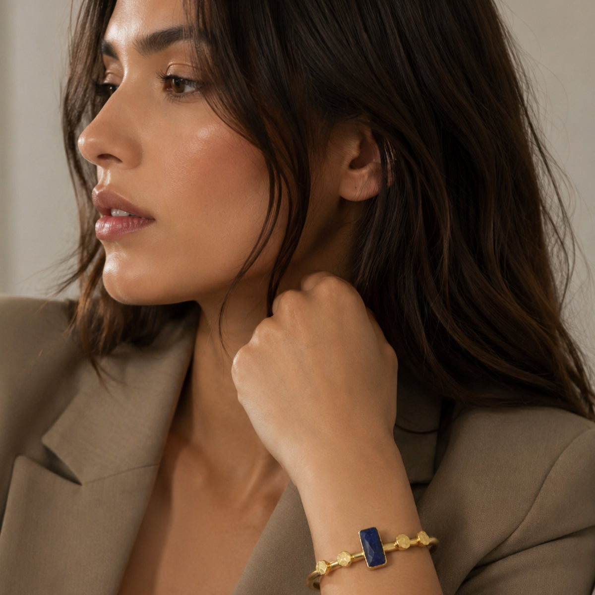 A close-up lifestyle shot of a woman wearing a gold bracelet with a rectangular blue lapis lazuli stone, paired with a beige blazer.