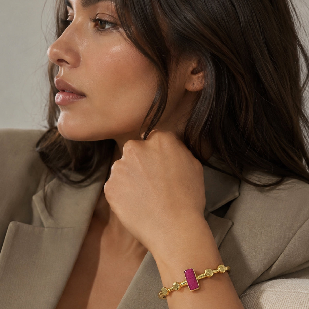 A close-up lifestyle shot of a woman wearing a gold bracelet with a rectangular magenta-pink ruby stone, posed in a beige blazer.