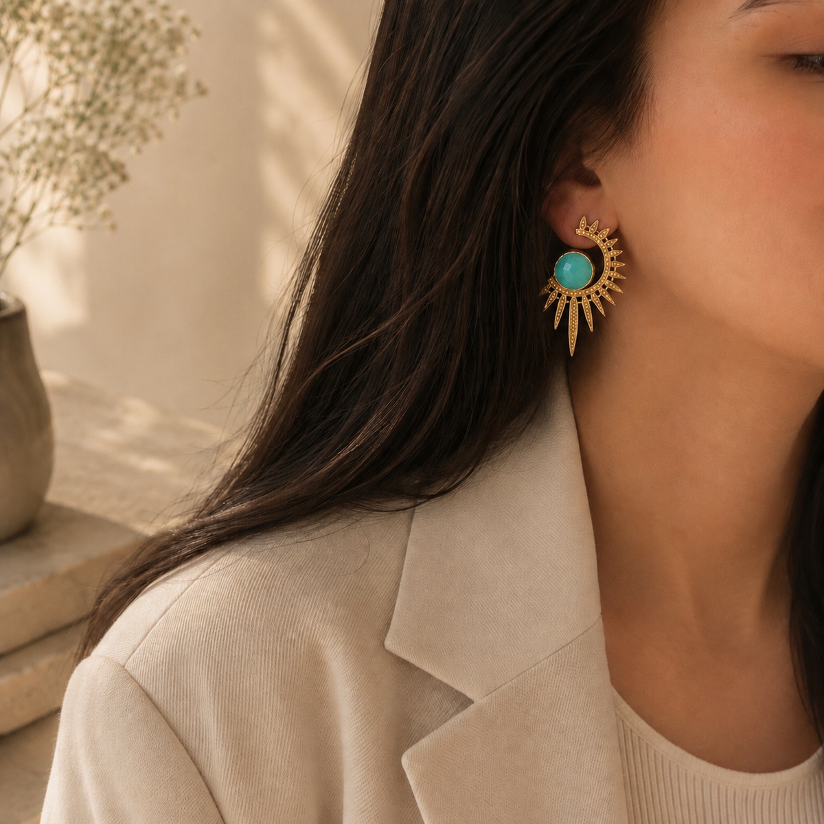 A close-up lifestyle shot of a woman wearing a gold crescent sunburst earring with a round aqua chalcedony stone, set against a warm beige background.