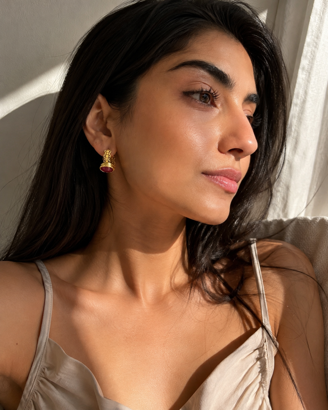 A close-up lifestyle shot of a woman with long dark hair wearing a small gold-plated hoop earring with a red ruby gemstone accent, set against a warm, sunlit neutral background.