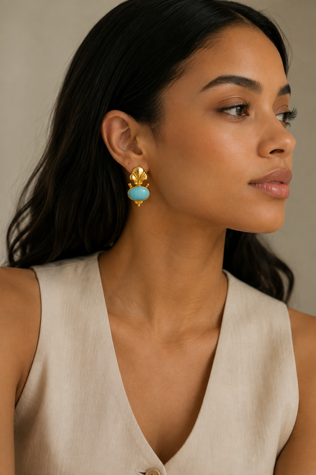 A close-up of a black woman wearing a pair of gold fleece de lis earrings with light blue turquoise oval stones, paired with a beige sleeveless top against a neutral beige background.