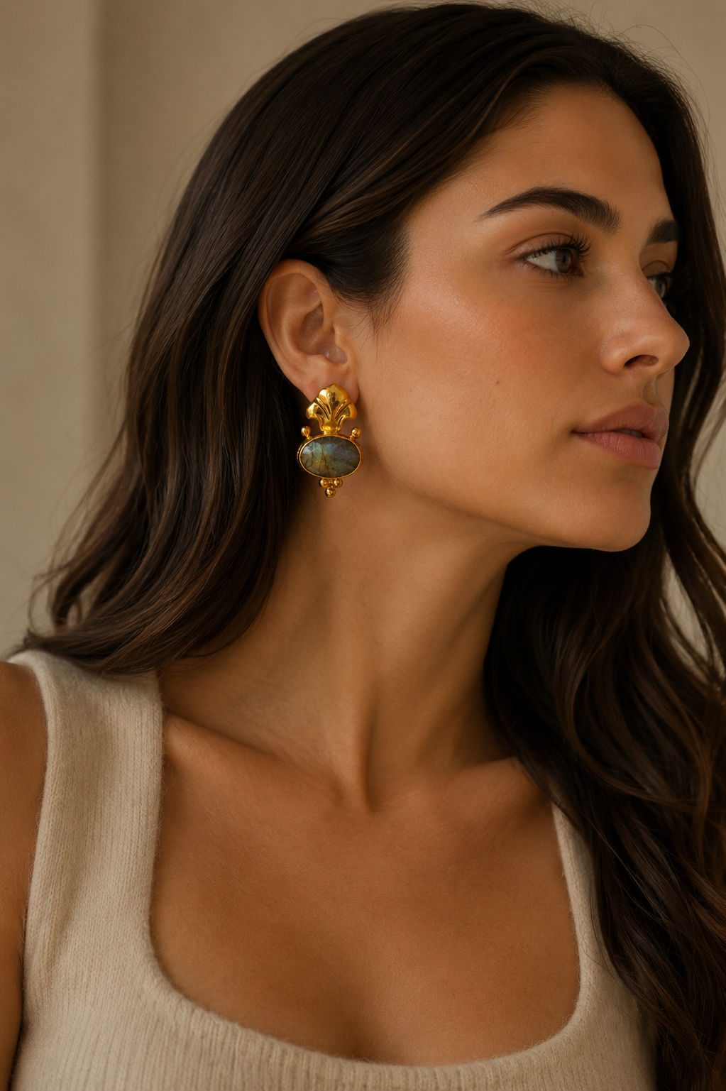 A close-up of a woman wearing a gold Fleur de lis statement earring with a round, iridescent gray-green labradorite stone, paired with a beige ribbed tank top.