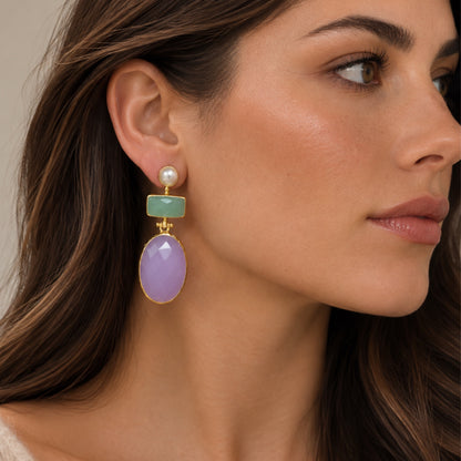 A close-up of a woman wearing a gold drop earring with a white pearl, a light green rectangular stone, and a large oval lavender-purple stone.