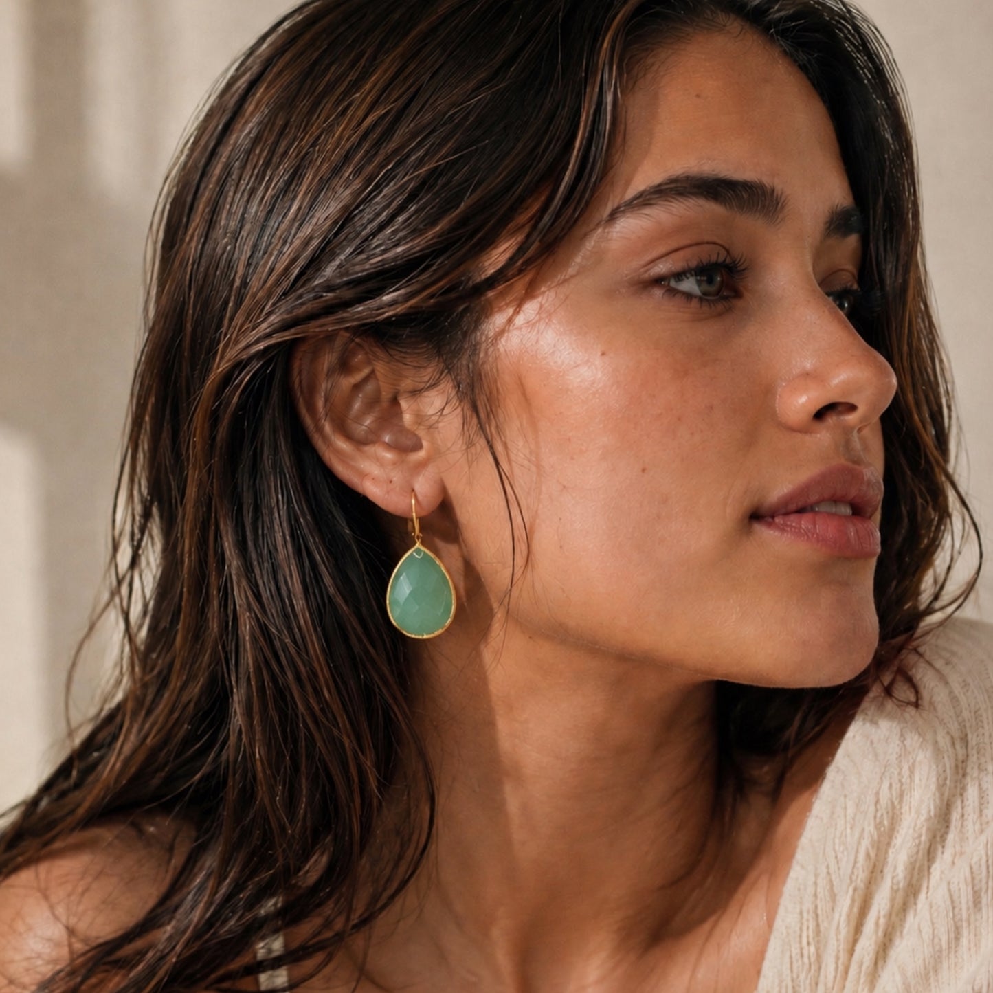 A close-up of a woman wearing a gold drop earring with an aqua chalcedonh teardrop-shaped gemstone, shown in profile against a warm neutral background.
