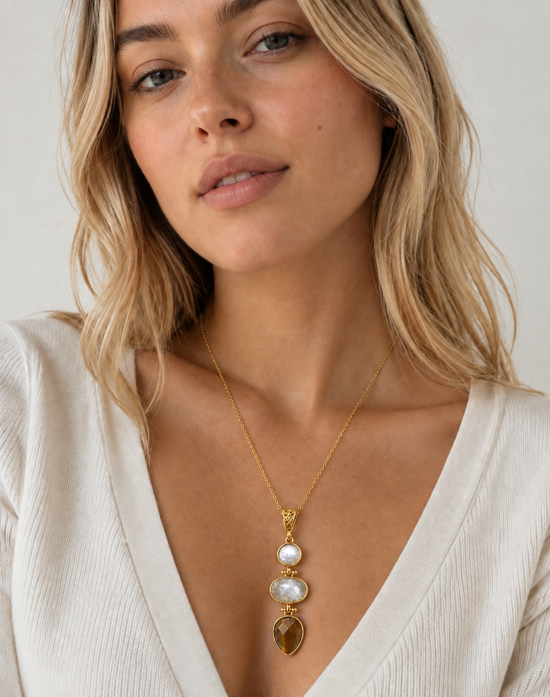 A close-up of a woman wearing a gold necklace with a vertical drop pendant featuring flat pearl, moonstone and a teardrop-shaped yellow agate gemstone, paired with a cream-colored ribbed top
