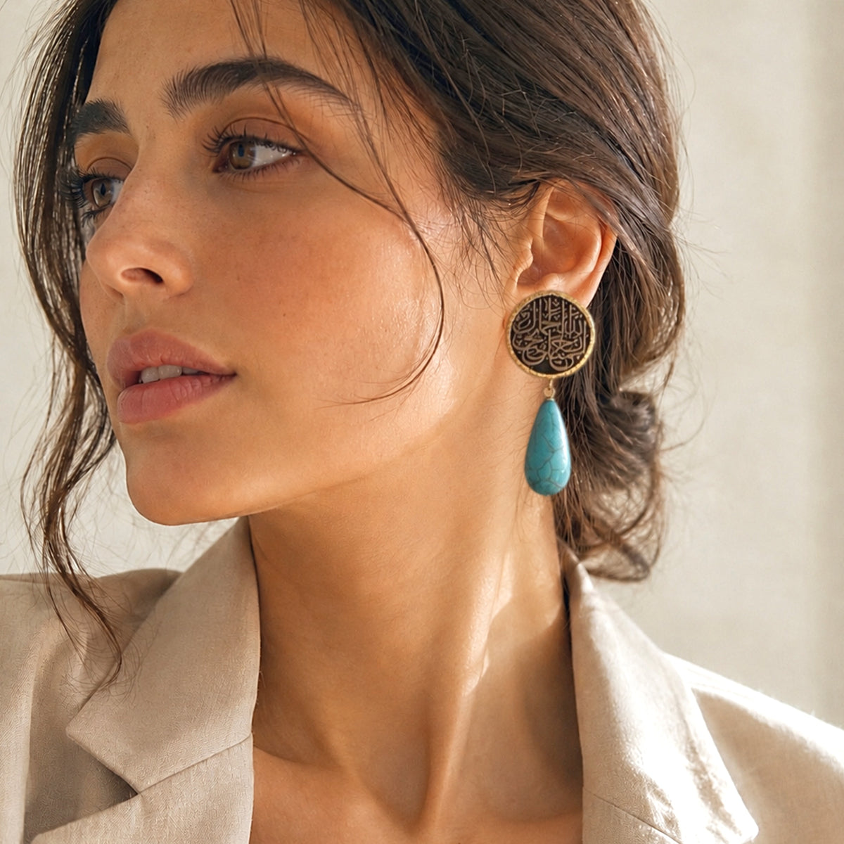 A close up of a woman wearing a gold plated Arabic calligraphy dangle earring with a circular filigree top and a turquoise teardrop drop paired with beige blazer