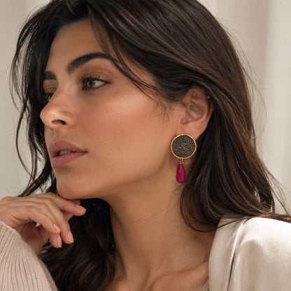 A close-up of a woman wearing a gold-plated dangle earring with a textured circular disc with Arabic calligraphy  and a dangling ruby on a soft beige background.
