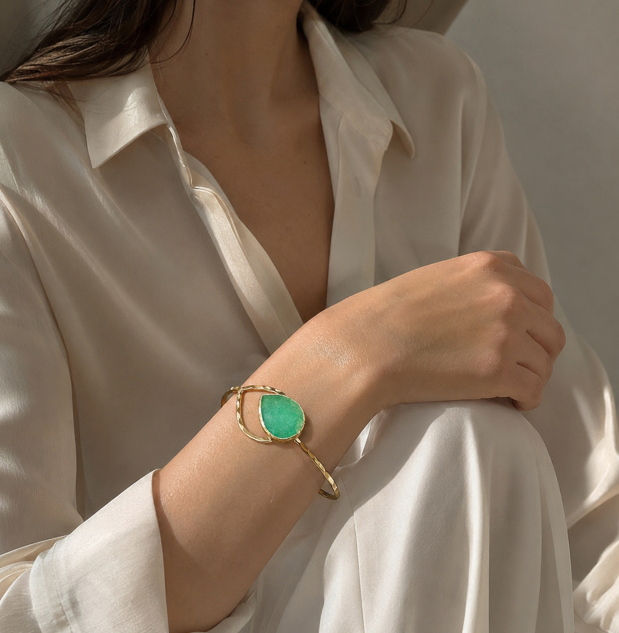 A close-up of a woman wearing a gold-plated green quartz cuff bracelet with a large green teardrop-shaped stone, styled with a white satin blouse.