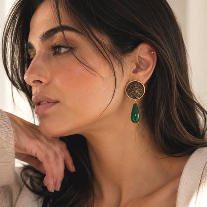 A close-up of a woman wearing a gold-toned drop earring with a gold and black circular textured medallion with Arabic calligraphy and a dangling green jade teardrop, against a soft neutral background.