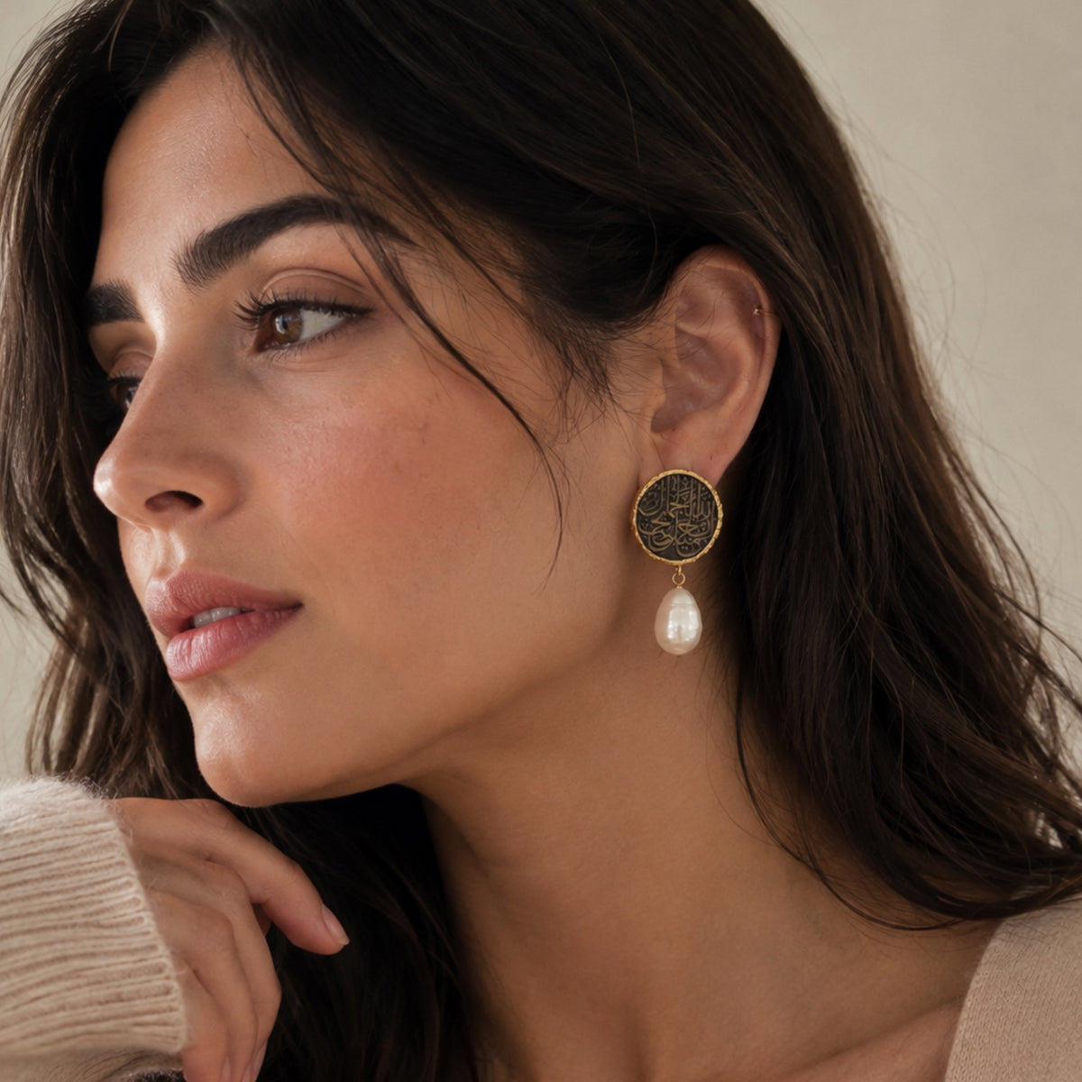 A close-up of a woman wearing a gold-toned drop earring with a gold plated Arabic calligraphy circular earring with dangling white baroque pearl, against a soft beige background.
