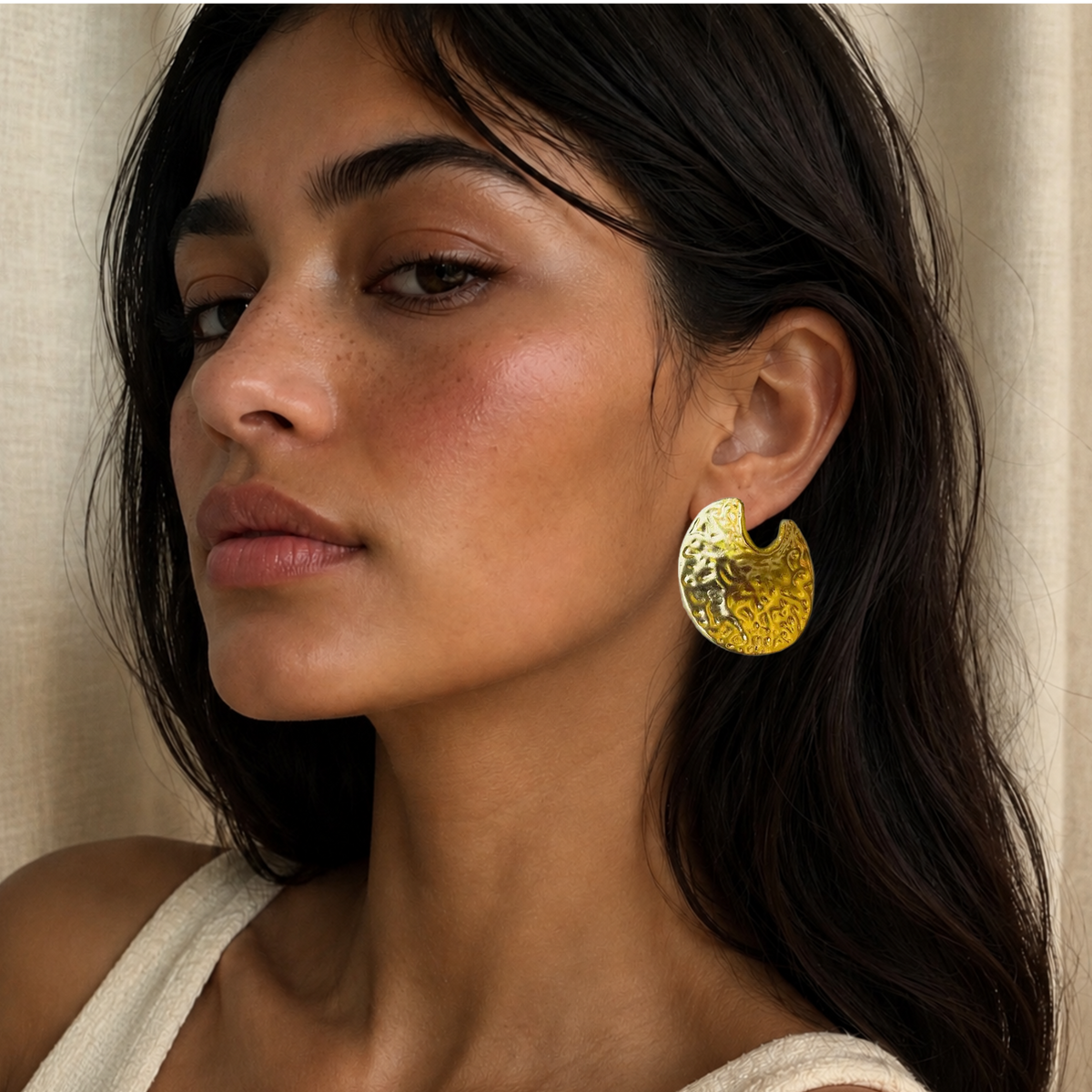 A close-up of a woman wearing a large, textured contemporary gold crescent-shaped earring, set against a soft beige curtain background.