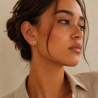 A close-up of a woman wearing a small gold evil eye-shaped stud earring with zircon center , shown in profile against a warm beige background.
