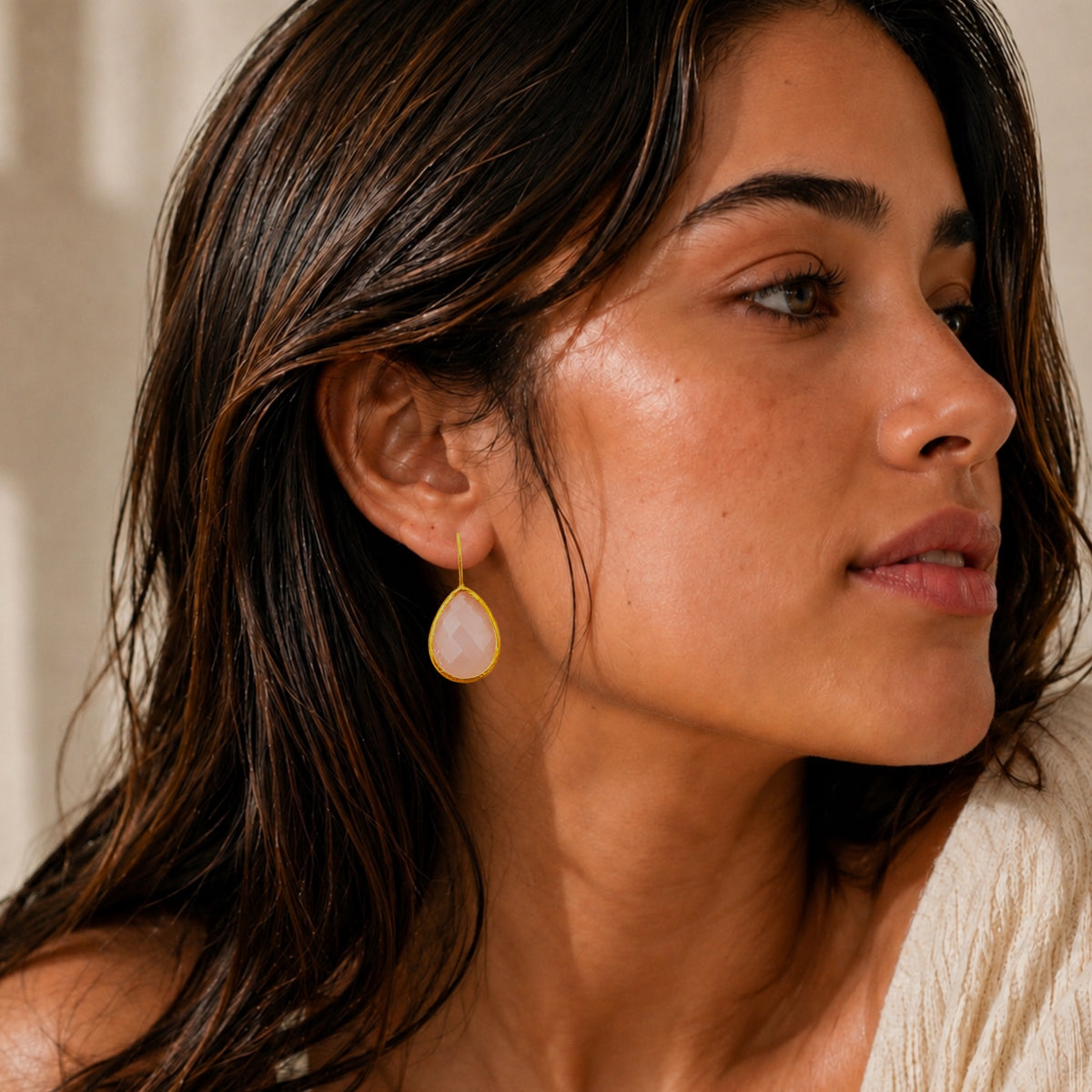 A close-up of a woman wearing a teardrop-shaped drop earring with a gold-toned rim and a pink rose quartz center shown against her ear and side profile.