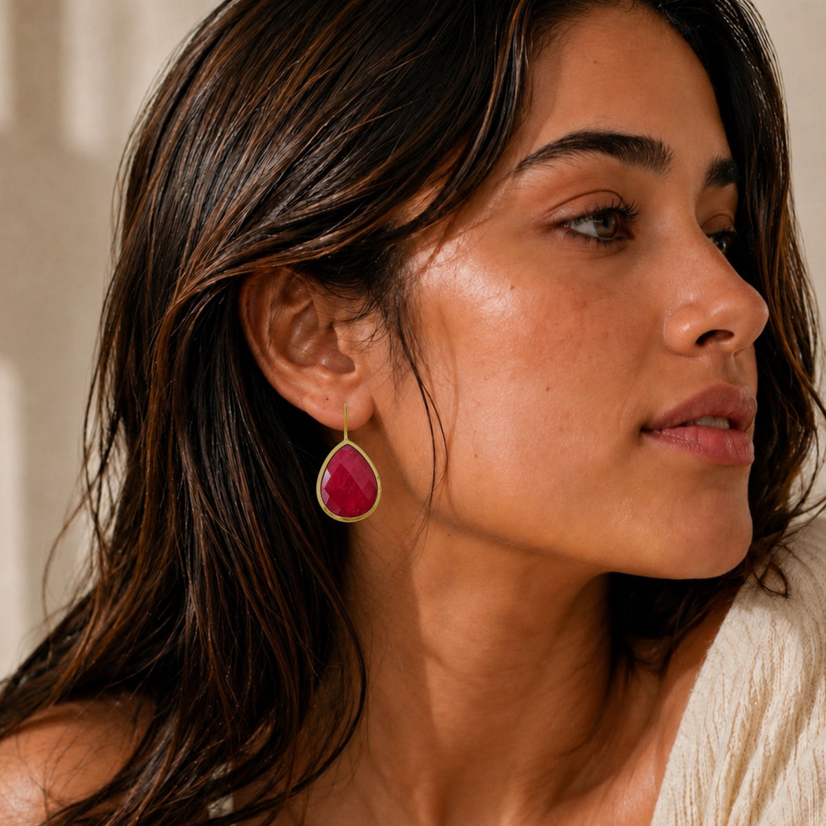 A close-up of a woman wearing an off-shoulder top and a teardrop-shaped ruby gemstone dangle earring with a gold-toned bezel setting.