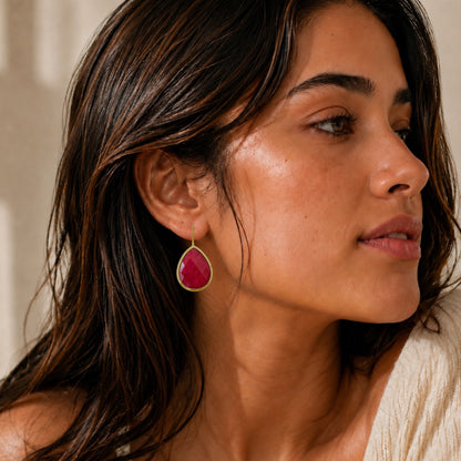 A close-up of a woman wearing an off-shoulder top and a teardrop-shaped ruby gemstone dangle earring with a gold-toned bezel setting.