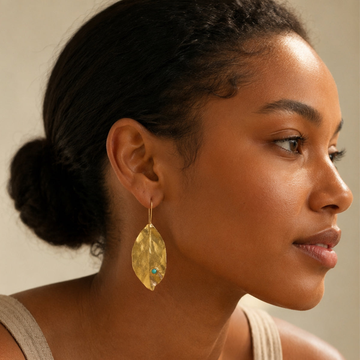 A close-up side profile of a woman wearing a textured gold leaf-shaped dangle earring with a small turquoise stone accent.