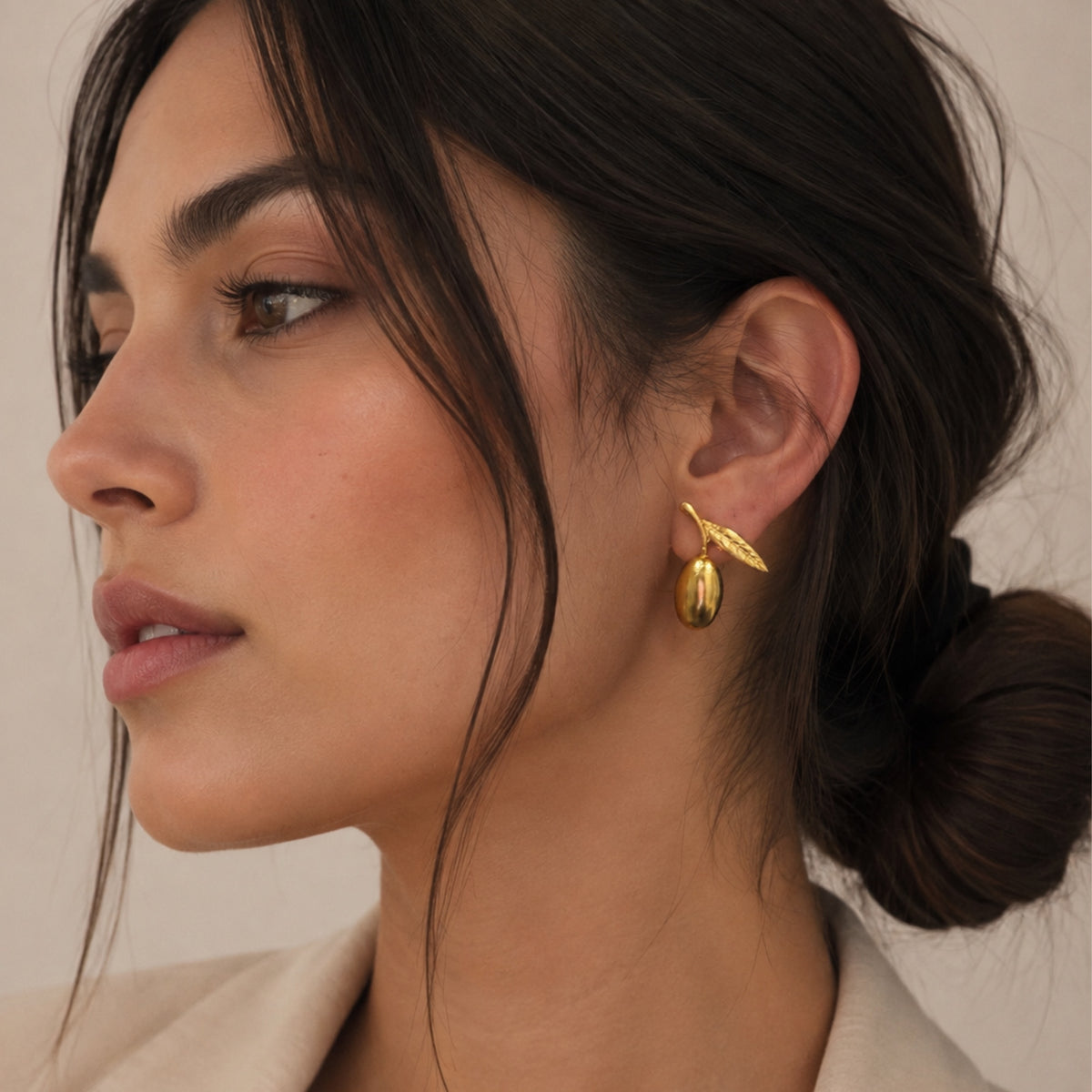 A close-up side profile of a woman with dark hair in a low bun, wearing a small gold olive shaped earring with leaf