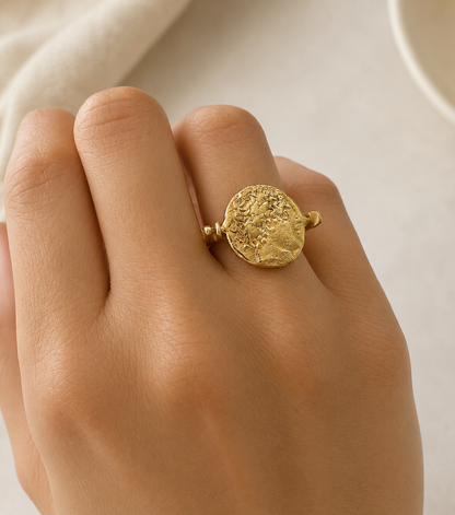 A gold coin-style flip ring with a textured, embossed circular face, shown worn on a person's finger against a soft, light beige background.