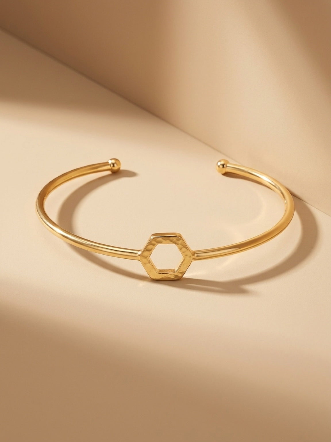A gold-plated brass cuff bracelet with a central open hexagon design shown on a plain beige background.