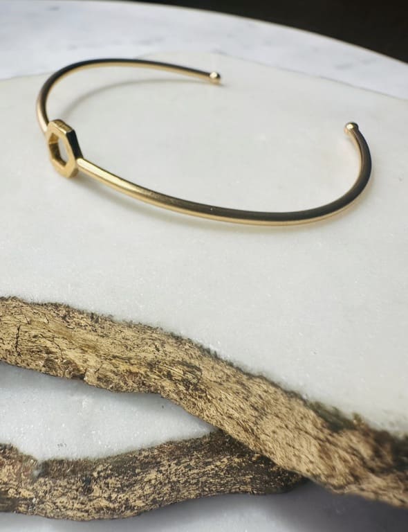 A gold-plated brass cuff bracelet with a central open hexagon design shown on a white marble background.