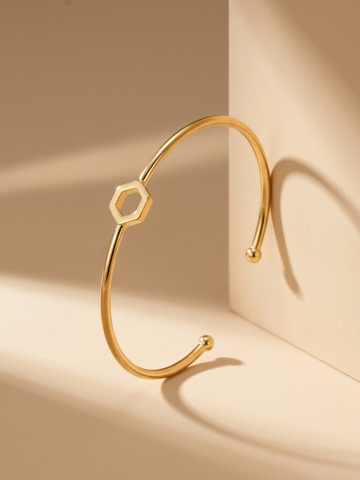 A gold-plated brass cuff bracelet with a central open hexagon design upright on a plain beige background.
