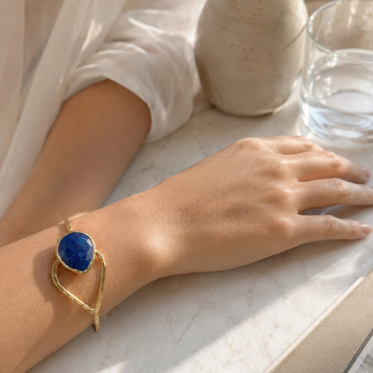 A gold-plated hammer-finished bracelet with a teardrop shaped blue lapis lazuli stone, worn on a person's wrist resting on a light marble surface.