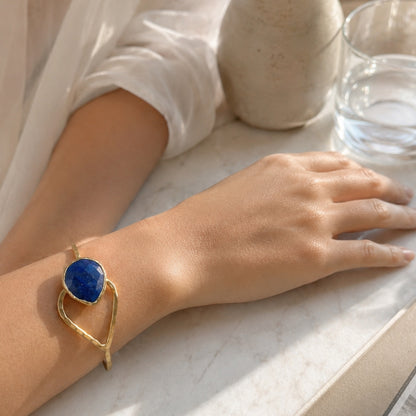 A gold-plated hammer-finished bracelet with a teardrop shaped blue lapis lazuli stone, worn on a person's wrist resting on a light marble surface.