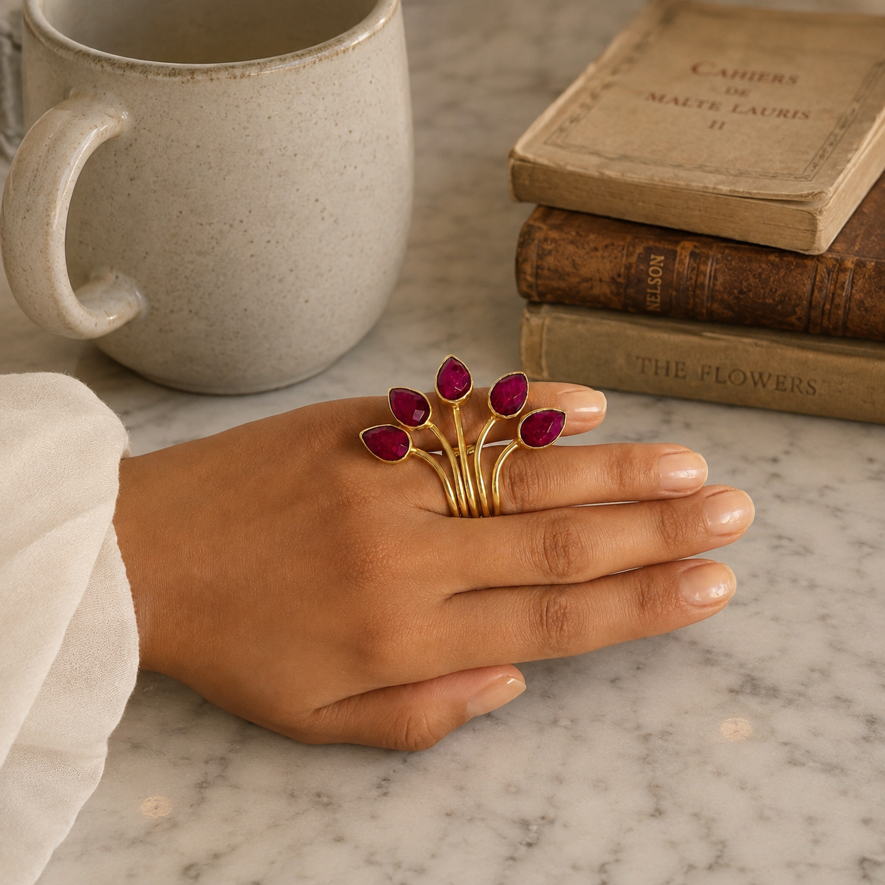 A gold ring with five leaf-shaped settings set with deep red ruby gemstones, worn on a person's hand resting on a white marble surface next to a ceramic mug and vintage books.