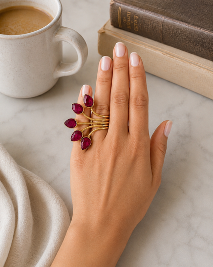 A gold ring with five teardrop-shaped deep red ruby gemstones worn on a hand, styled on a marble surface with a coffee mug and books in the background.