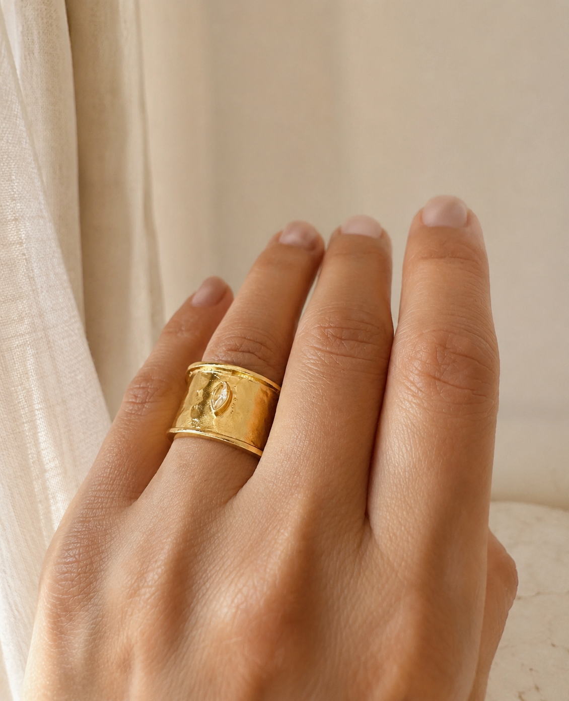 A close-up of a hand wearing a wide gold-plated band ring with a textured, hammered finish and a small central zircon detail, set against a soft beige background.