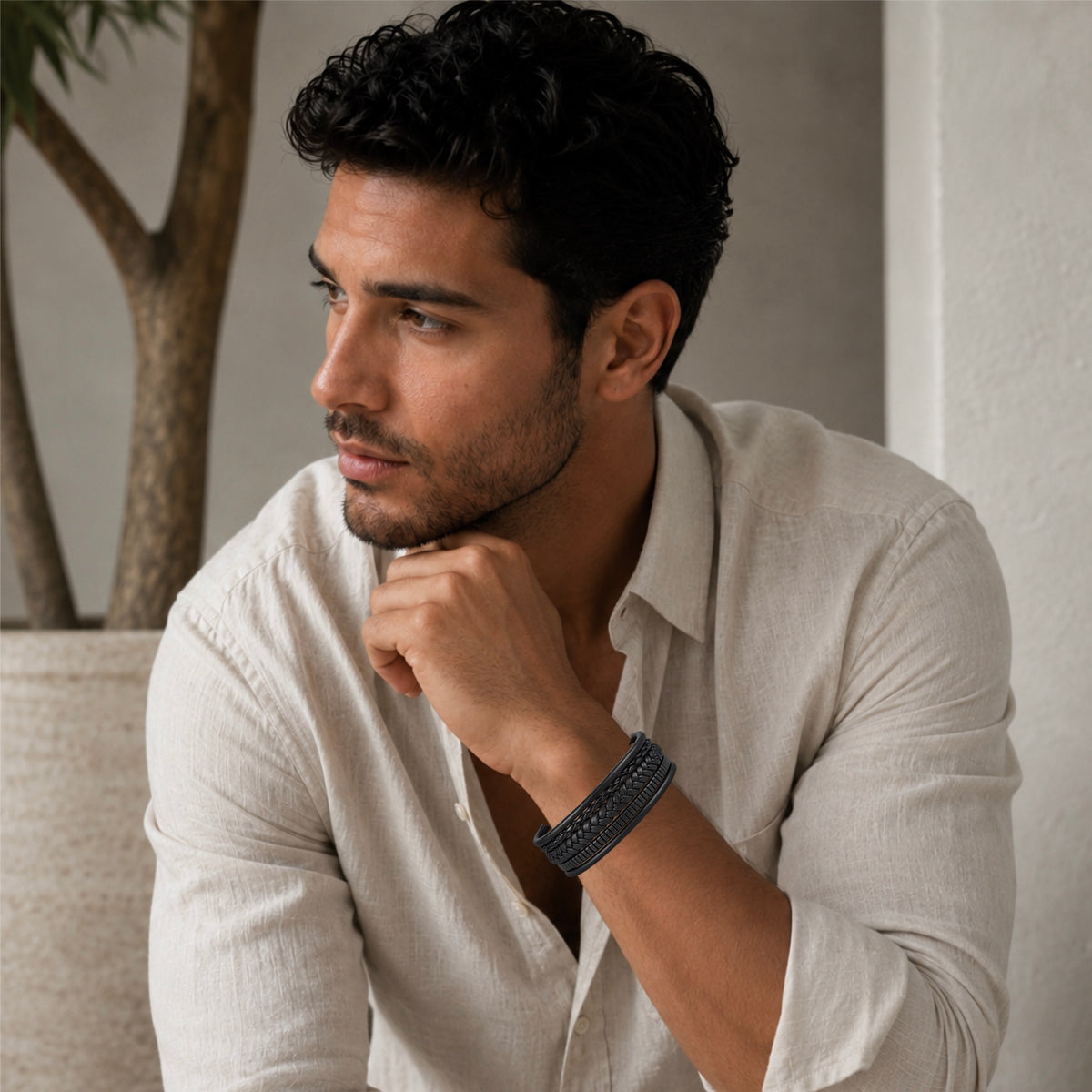 A man wearing a boho chick black woven leather bracelet, posing in a light beige linen shirt in a softly lit indoor setting.