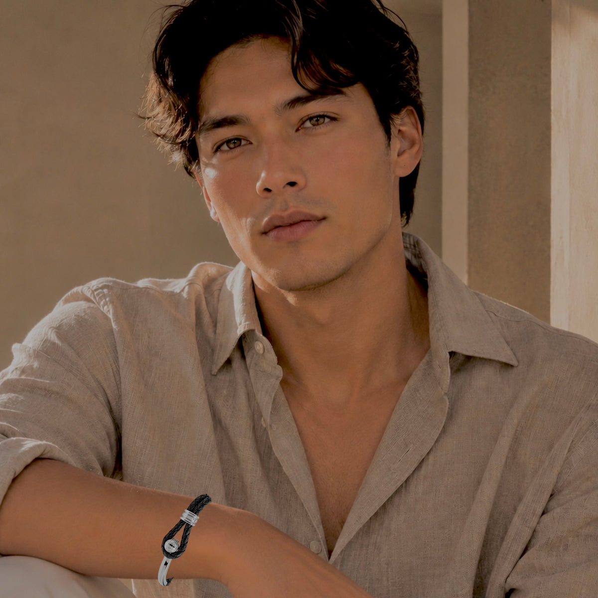 A man wearing a light beige linen shirt and a black and white braided bracelet with silver hardware, posed in a warm, neutral-toned indoor setting.