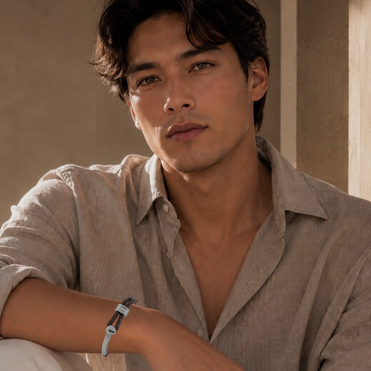A man wearing a light beige linen shirt and a braided brown leather bracelet with a silver buckle, posed in a warm, sunlit indoor setting.
