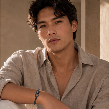 A man wearing a light beige linen shirt and a matte black leather bracelet with matte black hardware, in a warm, sunlit indoor setting.